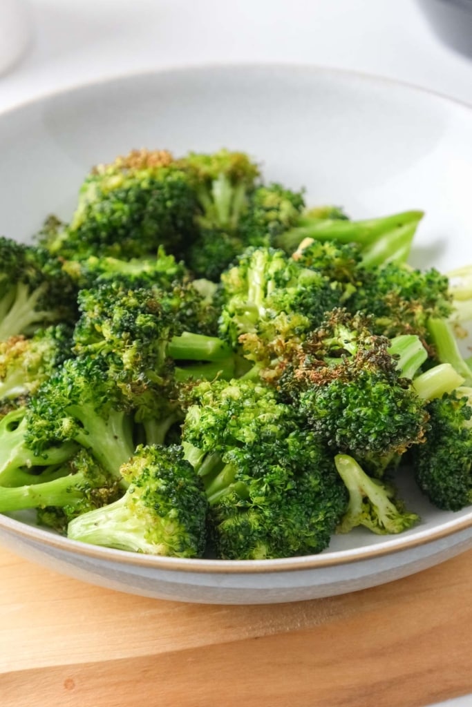fried broccoli florets in white bowl on wooden board on white counter.