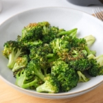 green cooked broccoli in white bowl on wooden board with black air fryer handle behind.
