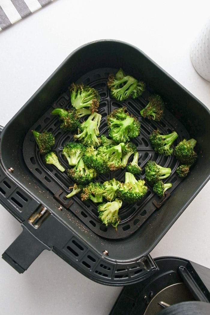 broccoli florets in black air fryer tray on white counter top.