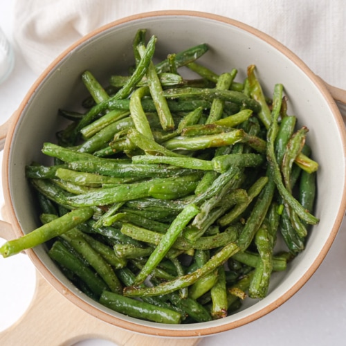 crispy green beans in serving dish with one hanging out sitting on wooden board on counter.