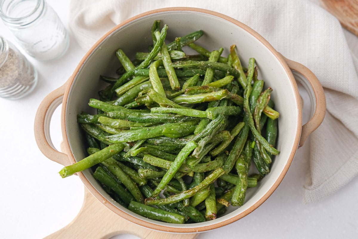 crispy green beans in serving dish with one hanging out sitting on wooden board on counter.