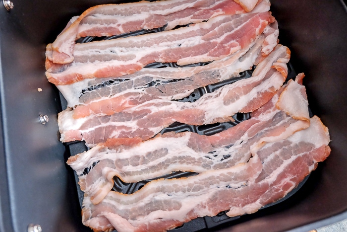 slices of raw bacon laid on black air fryer tray