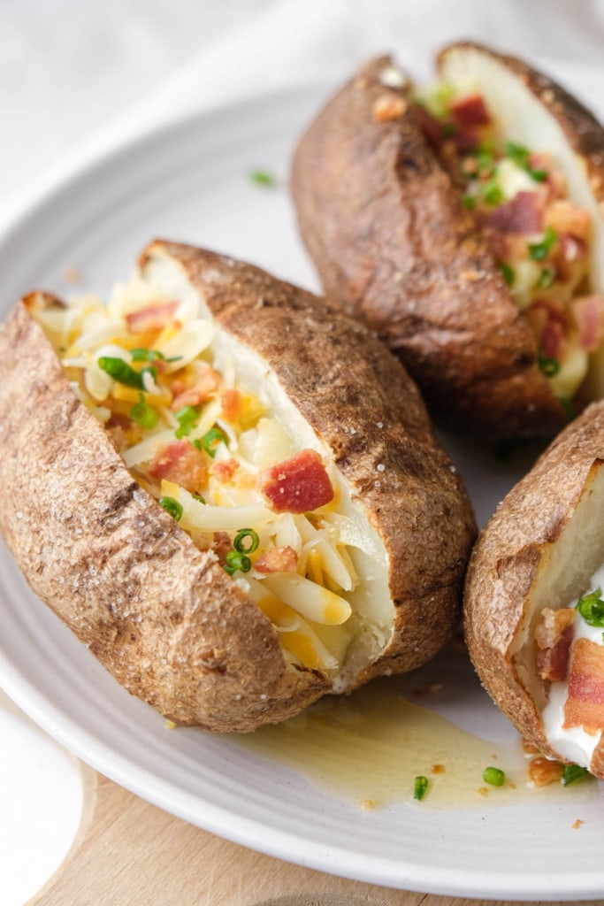 baked potatoes cut in half with bacon butter and cheese in the middle sitting on white plate.