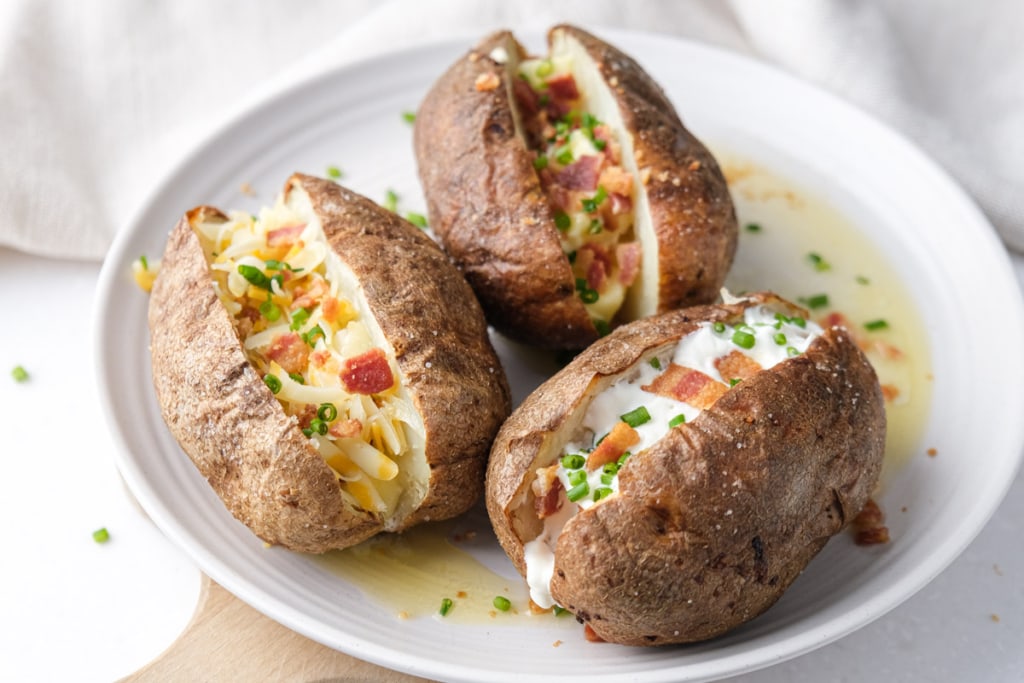three fully loaded baked potatoes on white plate sitting on wooden board.