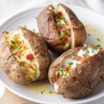 three fully loaded baked potatoes on white plate sitting on wooden board.