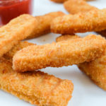 crispy breaded fish sticks on white plate with ketchup behind