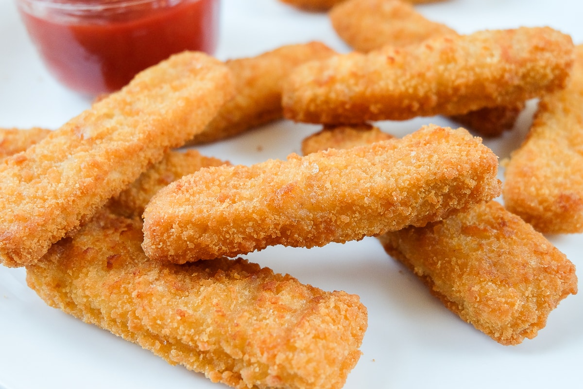 crispy breaded fish sticks on white plate with ketchup behind