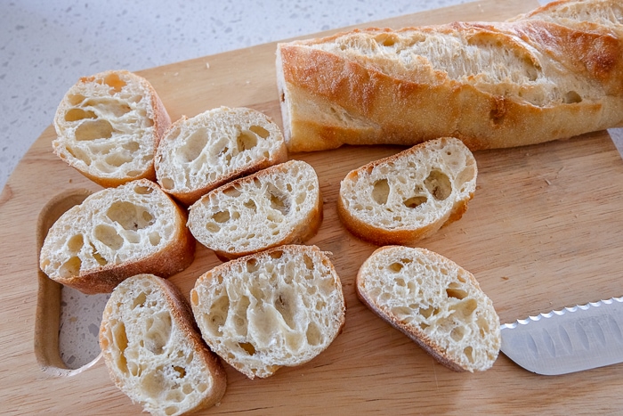 slices of french baguette on wooden cutting board with loaf behind