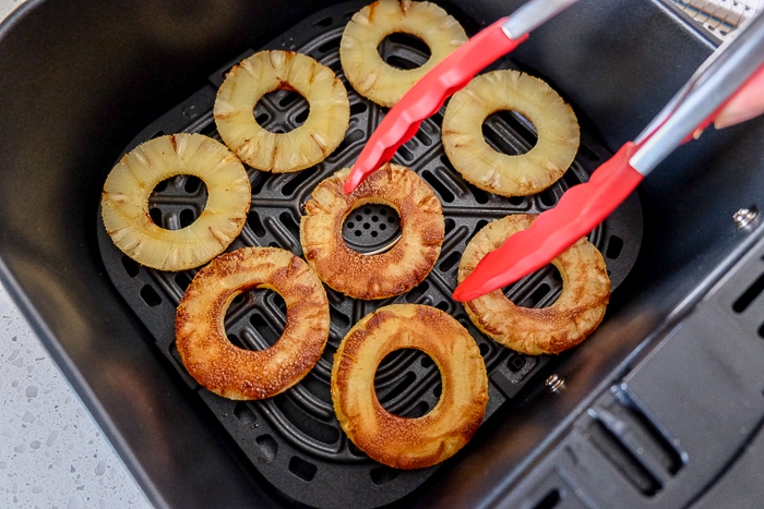 red tongs flipping pineapple slices in black air fryer tray