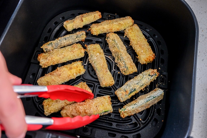 red tongs flipping breaded zucchini fry in air fryer tray