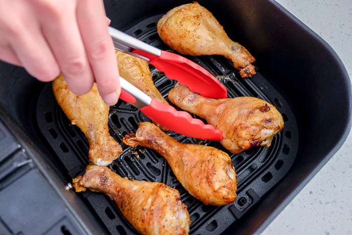 chicken drumsticks in air fryer flipping with red tongs