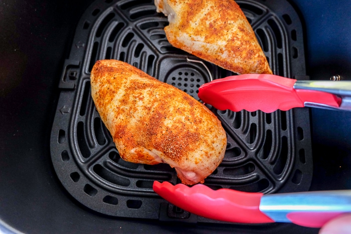 flipping chicken breast in black air fryer tray with red tongs
