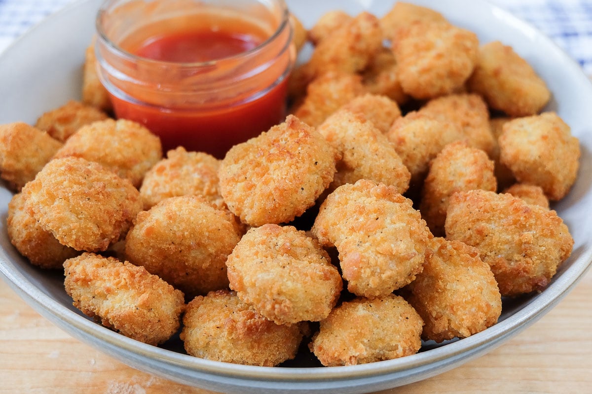 bowl of crispy breaded popcorn chicken with red dipping sauce behind all on wooden board.