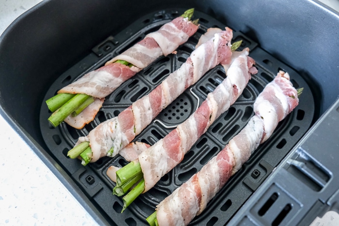 raw bacon wrapped asparagus in black air fryer tray on counter