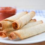 cooked taquitos on white plate with dipping salsa behind