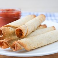 cooked taquitos on white plate with dipping salsa behind