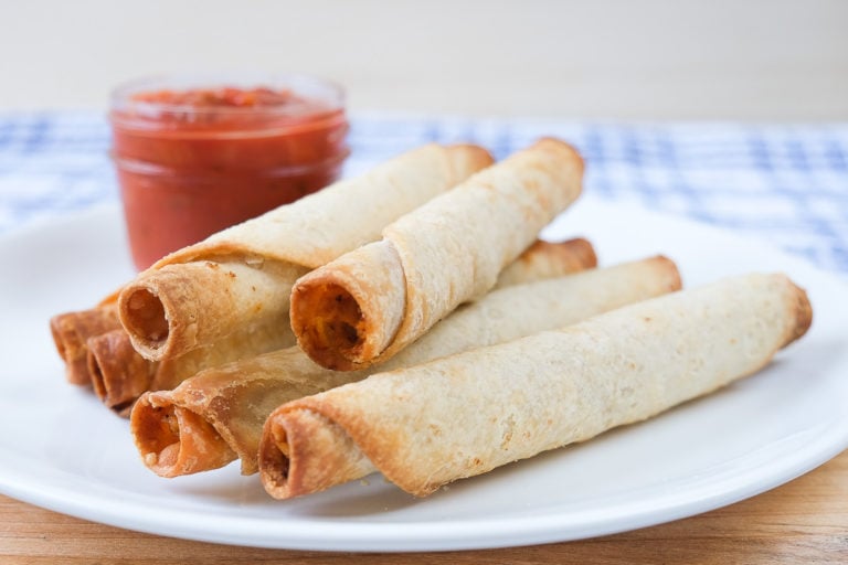 cooked taquitos on white plate with dipping salsa behind