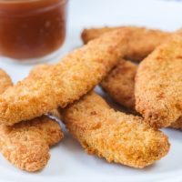 crispy breaded chicken strips on white plate with plum sauce behind