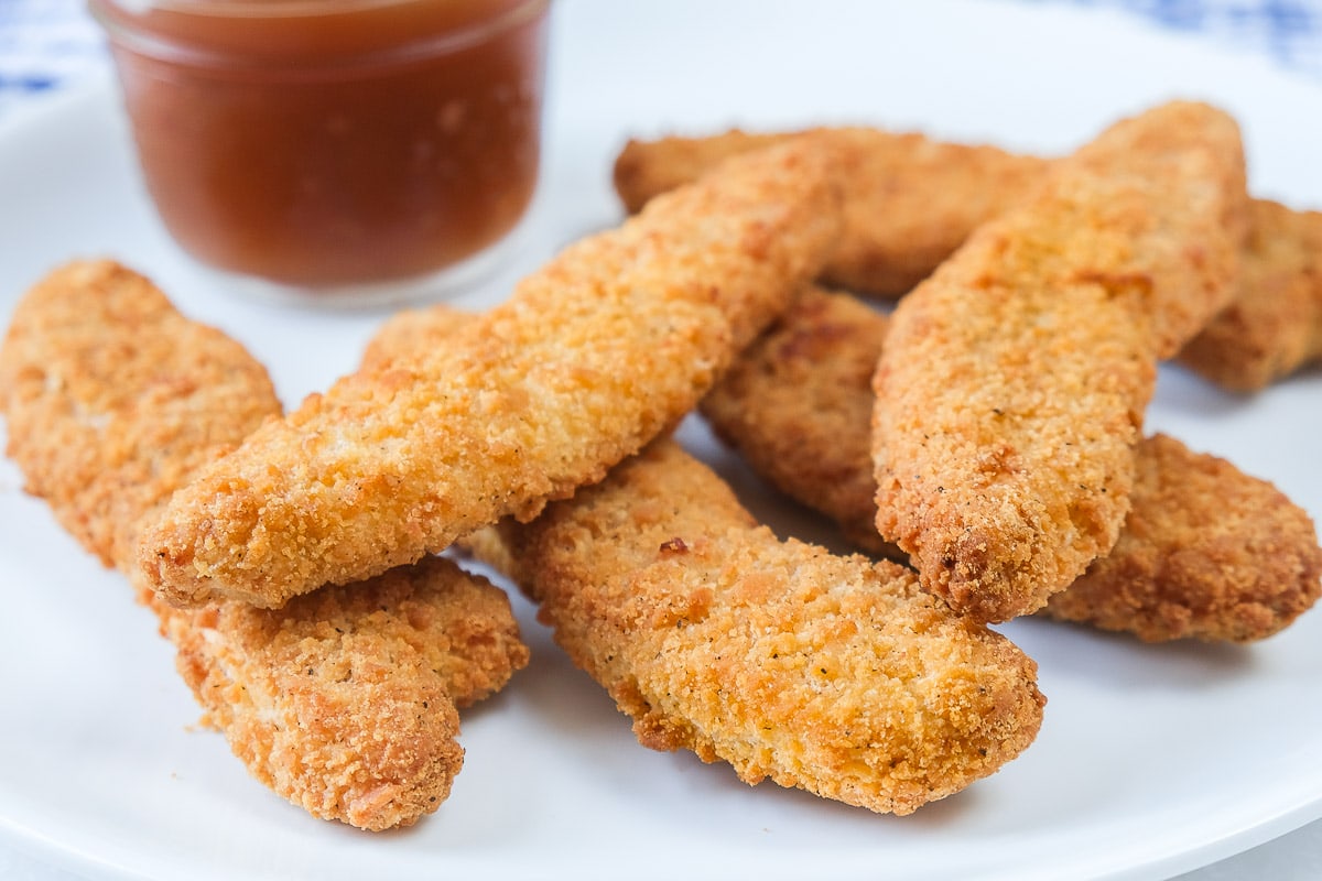 crispy breaded chicken strips on white plate with plum sauce behind