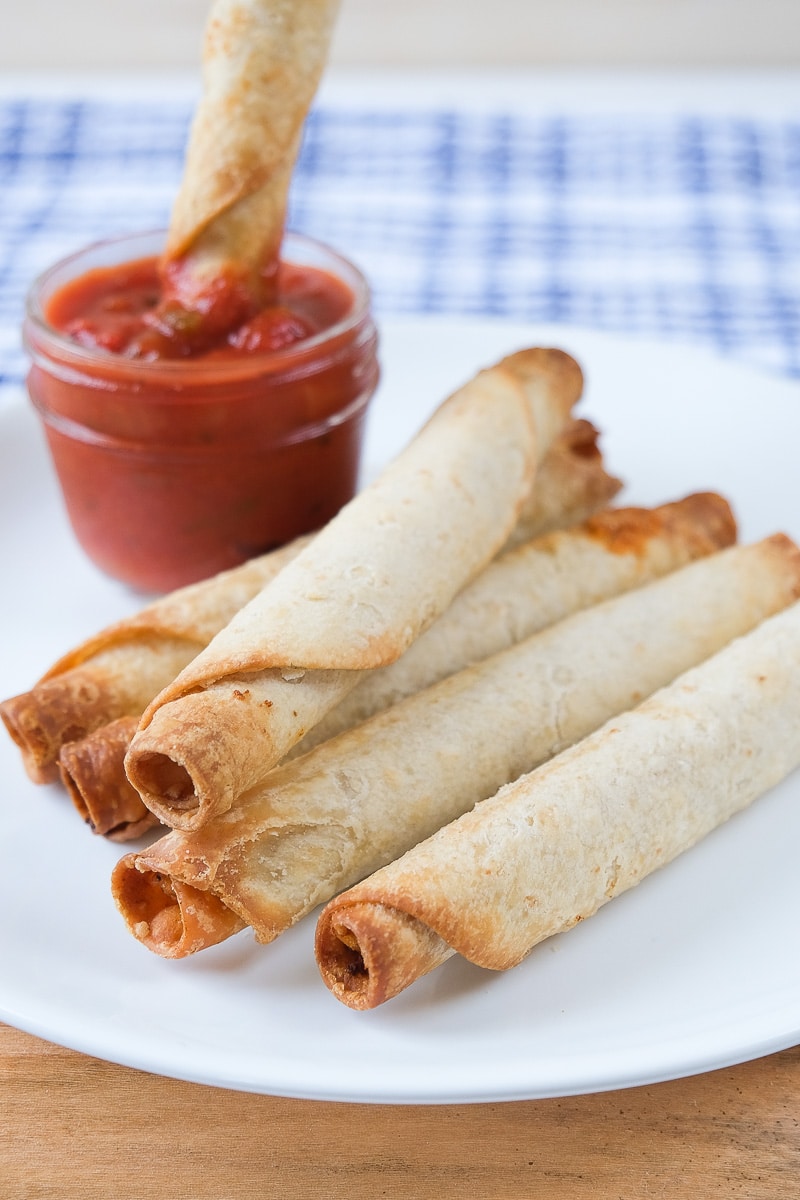 crispy taquitos in white plate with one dipping in salsa behind