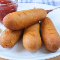 cooked corn dogs on white plate with ketchup behind