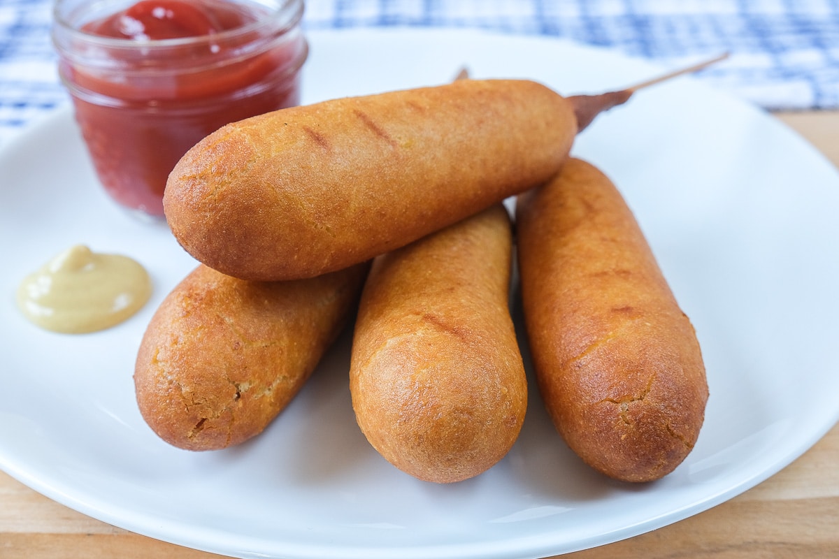 cooked corn dogs on white plate with ketchup behind