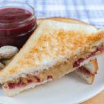 half of a peanut butter and jelly sandwich on plate with jam and peanuts behind