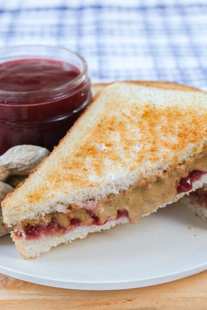 peanut butter and jelly sandwich on white plate with jam behind