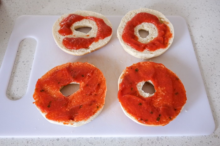 red tomato sauce spread on pizza bagels on white cutting board