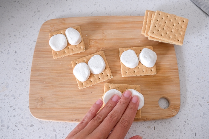 hand pressing down marshmallows onto graham crackers on wooden board