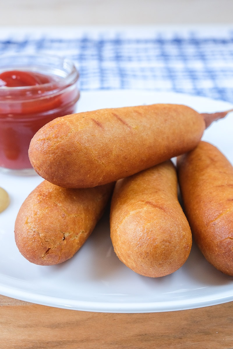brown corn dogs on white plate with ketchup behind