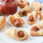 pigs in a blanket on white plate with dip bowl behind