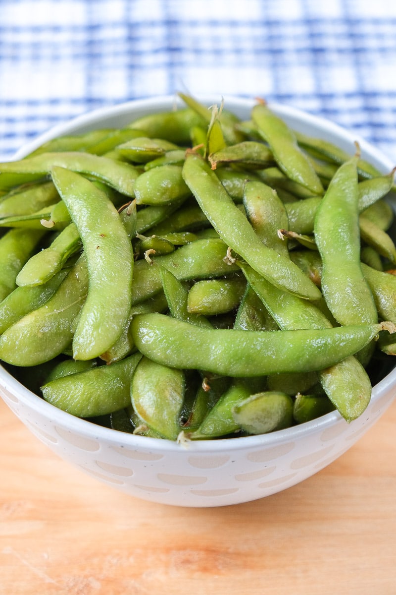 green edamame in bowl on wooden board with blue cloth behind