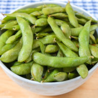 bowl of green edamame on wooden board with blue cloth behind