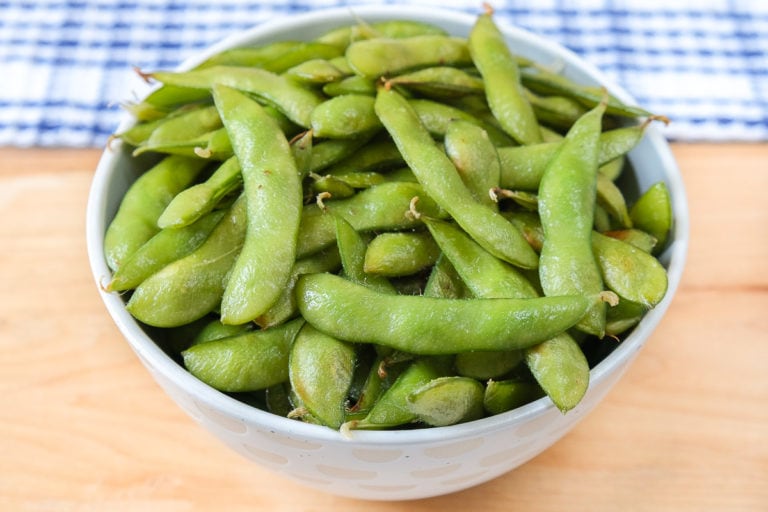 bowl of green edamame on wooden board with blue cloth behind