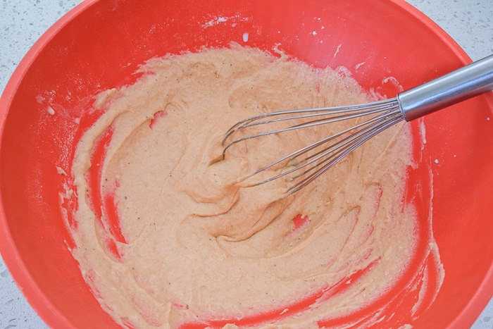 red mixing bowl of wet batter with metal whisk stirring