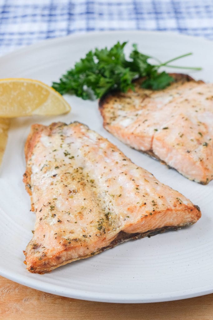 cooked salmon fillets on white plate with lemons and parsley behind