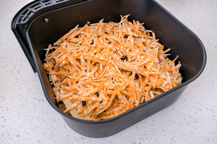 shredded cheese covering tortilla chips in black air fryer tray on white counter
