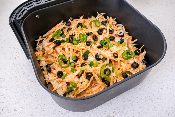 jalapeno peppers on uncooked nachos in black air fryer tray on white counter