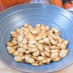 roasted pumpkin seeds in blue bowl on wooden board with pumpkin behind