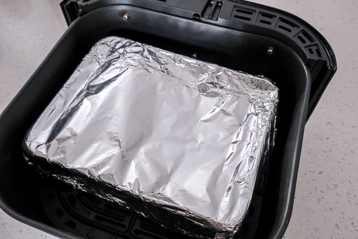 glass dish of apple crisp covered with tin foil in black air fryer tray on counter