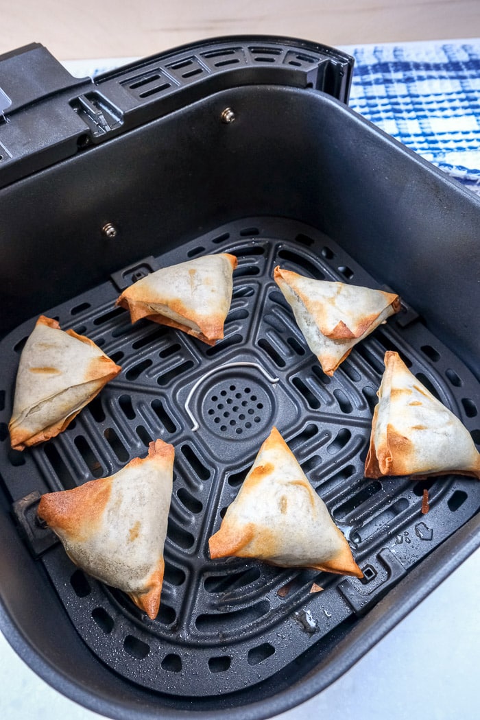 crispy samosas in black air fryer tray on counter with blue cloth behind