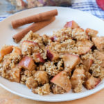 white plate on wood board with apple crisp on top with cinnamon sticks behind
