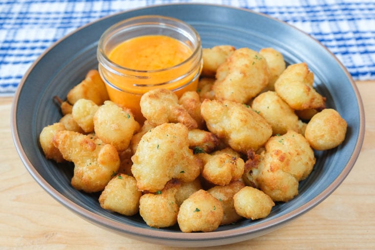 buffalo cauliflower bites in blue bowl on wooden board with buffalo sauce beside