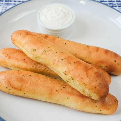 golden brown breadsticks on large plate on wooden board with dip behind