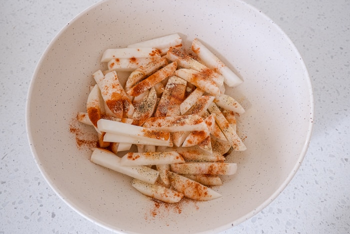 white bowl on white counter with raw turnip fries and seasonings on top.