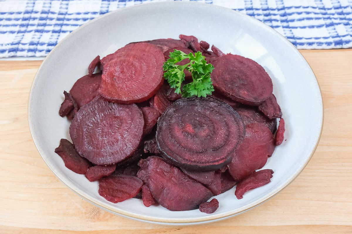 cooked sliced beets in bowl on wooden board with parsley on top.