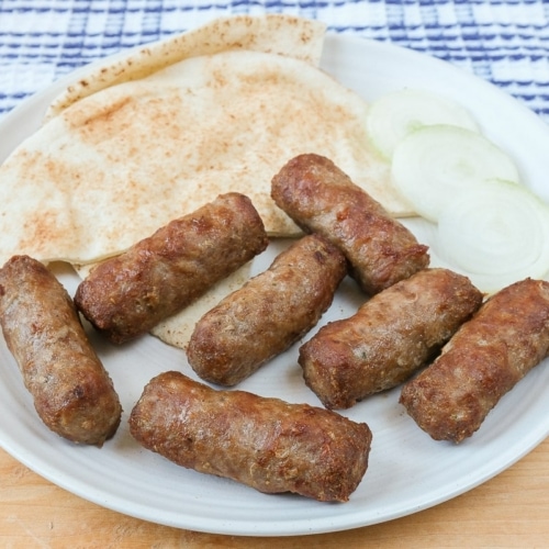 grilled sausages on white plate on wood with pita and sliced onion behind.