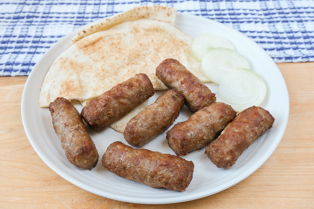 grilled sausages on white plate on wood with pita and sliced onion behind.