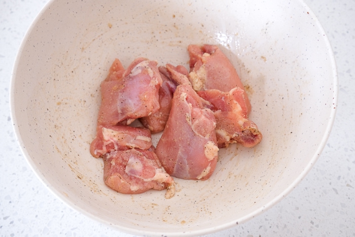raw chicken thighs coated in oil and spices in white mixing bowl.